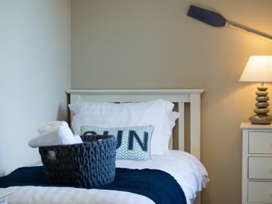 A bedroom with a single bed with white and blue bedding a basket with towels on the bed a bedside table with a lamp and a blue paddle on the wall at Cargoes in Craster