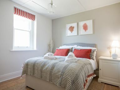 A bedroom with a bed with red pillows and a patterned blanket a window with striped blind and a bedside table with a lamp at Cargoes in Craster