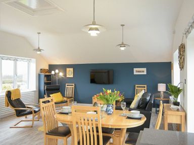 A dining area with a wooden table set with dishes and flowers and a living area with black chairs and a wall mounted tv at Viewpoint in Alnmouth