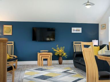 A living room with a blue accent wall a mounted television yellow and gray area rug wooden chairs a black sofa and a vase with yellow flowers at Viewpoint in Alnmouth