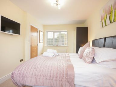 A bedroom with a bed covered in a pink quilt and pillows a TV mounted on the wall a window with blinds and a wardrobe at Viewpoint in Alnmouth