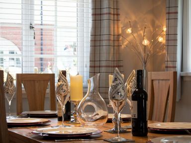 A dining table set with plates wine glasses napkins a decanter candles and wine bottles in a room with wooden chairs and a window at Castlewood Bamburgh