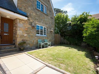 A stone house exterior with a wooden door, a window, two chairs, a small lawn, and surrounding trees at Castlewood in Bamburgh