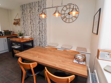 A dining area with a wooden table and four chairs near a corner bench with cushions and a large clock on the wall at Rosedale Lodge in Alnmouth