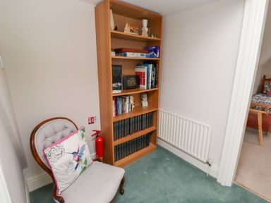 A bookshelf filled with books and decorative items next to a chair with a cushion and a fire extinguisher on a green carpet at Rosedale Lodge in Alnmouth