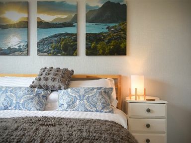 A bed with blue patterned pillows and a textured gray pillow a bedside table with a lamp and a triptych landscape artwork on the wall at Spuggies Rest in Embleton