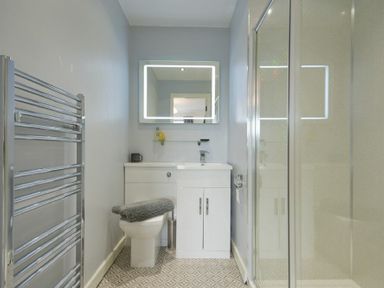 A bathroom with a toilet, vanity with sink, illuminated mirror, glass shower enclosure, and a wall-mounted towel radiator at Spuggies Rest in Embleton