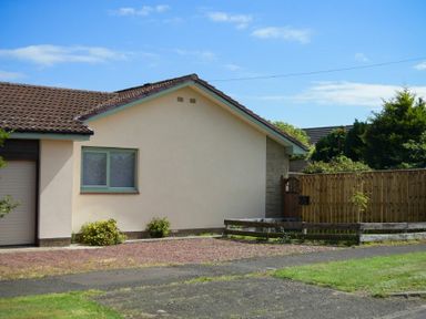 The side of a single-story house with a window a garage door gravel driveway wooden fence and some bushes with a blue sky in the background at Spuggies Rest in Embleton