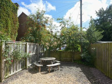 A fenced garden area with a round wooden picnic table and benches on gravel at Stewart House in Wooler