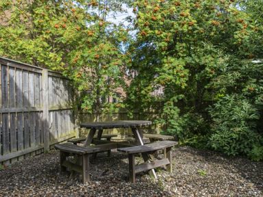 A wooden picnic table with attached benches in a garden with gravel ground and trees at Stewart House in Wooler