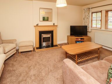 A living room with a beige armchair, ottoman, coffee table, TV on a wooden cabinet, fireplace, and windows with curtains at Stewart House in Wooler