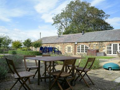 An outdoor patio area with a wooden table and six chairs near a stone building with arched windows and doors at The Warren in Bamburgh