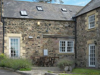 An outdoor patio with wooden table and chairs next to a stone house with white framed windows and doors at The Warren in Bamburgh