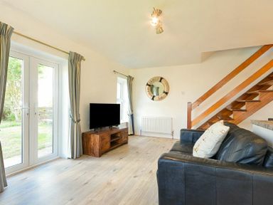 A living room with a black leather sofa a wooden TV stand with a TV curtains on windows and a wooden staircase at Coastal Plaice in Beadnell