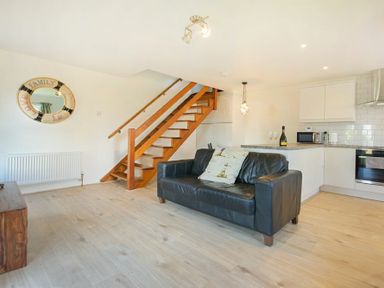 A living area with a black sofa two cushions wooden stairs and a kitchen with white cabinets at Coastal Plaice in Beadnell