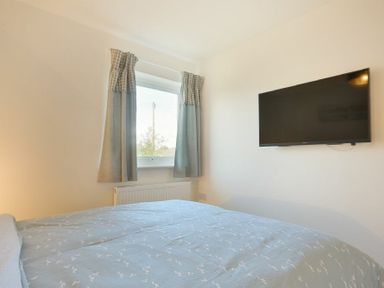 A bedroom with a bed covered in blue bedding a window with curtains and a wall-mounted television at Coastal Plaice in Beadnell