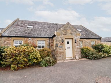 Stone house with white door and multiple windows surrounded by bushes at The Wynd in Bamburgh