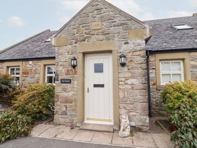 Stone house exterior with a white door and two outdoor wall lights plants on either side of entrance at The Wynd in Bamburgh