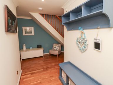 A hallway with wooden flooring a blue shelf a white bench a chair under stairs and decorations at The Wynd in Bamburgh