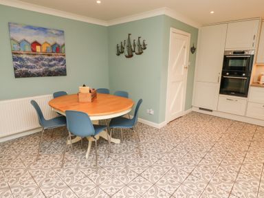 A dining area with a round wooden table and six blue chairs next to a kitchen with white cabinets at The Wynd in Bamburgh