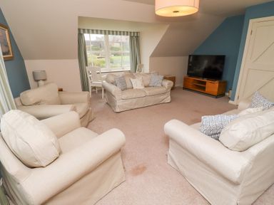 A living room with beige sofas a TV on a wooden stand and a window with chairs at The Wynd in Bamburgh
