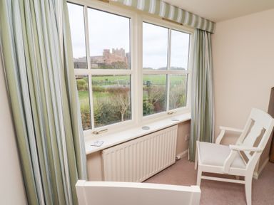 A room with a white wooden chair and striped curtains by a window overlooking a castle and green fields at The Wynd in Bamburgh