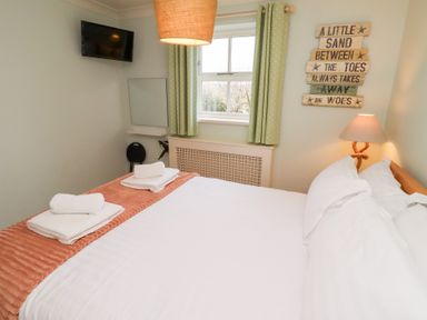 A bedroom with a bed with white linens and folded towels a wall-mounted tv a window with curtains and a lamp at The Wynd in Bamburgh