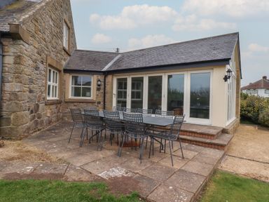 An outdoor patio with a long table and metal chairs beside a stone building with large windows at The Wynd in Bamburgh