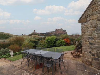 An outdoor patio with a metal table and chairs overlooking a garden and a distant castle at The Wynd in Bamburgh