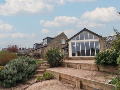 A stone house with a large window extension and garden steps surrounded by plants at The Wynd in Bamburgh