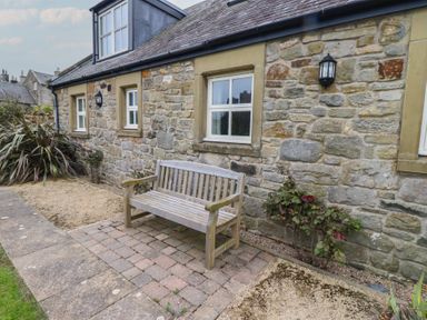 A stone house exterior with three windows a wooden bench and plants on a paved and gravel area at The Wynd in Bamburgh