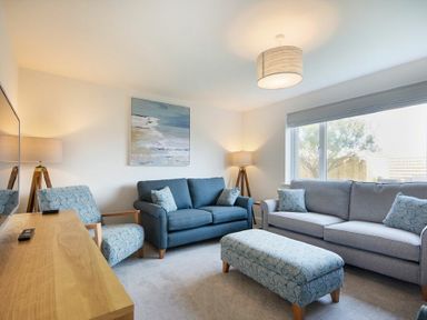 A living room with two sofas one armchair an ottoman wooden TV stand lamps and a large window at Luna Tide in Seahouses