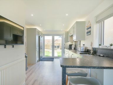 A kitchen with a wall-mounted television a countertop with two stools and glass doors leading outside at Luna Tide in Seahouses