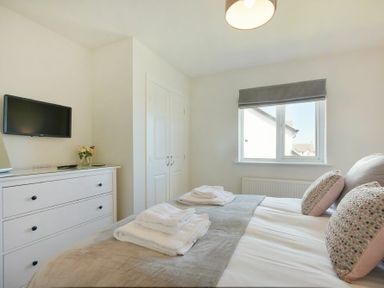 A bedroom with a double bed folded towels on the bed a white dresser with a vase of flowers a wall mounted tv a window and a closet with double doors at Luna Tide in Seahouses