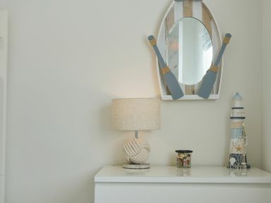 A white dresser with a rope lamp a jar decorative lighthouse and a mirror with two paddles on the wall at Luna Tide in Seahouses