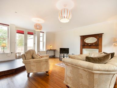 A living room with cream sofas a wooden floor a small TV an ornate wooden fireplace and large windows at The Old Concert Room in Alnmouth