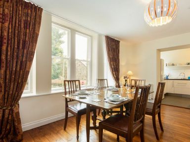 A dining room with a wooden table set for six near windows and an open doorway to a kitchen at The Old Concert Room in Alnmouth