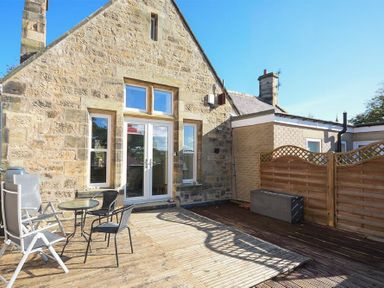 An outdoor wooden deck with a small round table and chairs next to a stone building with glass doors and windows at The Old Concert Room in Alnmouth