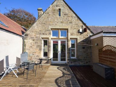 An outdoor patio with chairs and a table next to a stone house with windows and doors at The Old Concert Room in Alnmouth