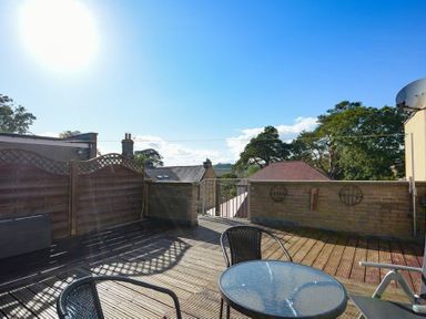 A wooden deck with a glass table and chairs overlooking rooftops and trees at The Old Concert Room in Alnmouth