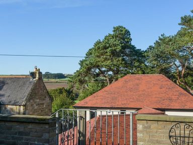 View of rooftops trees and fields behind a metal gate and stone wall at The Old Concert Room in Alnmouth