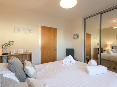 A bedroom with a bed with pillows and rolled towels a wooden door a chair a bookshelf and mirrored sliding closet doors at Coast View in Beadnell