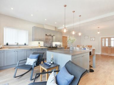 A kitchen with gray cabinets a cooking range an island with pendant lights and black chairs at Bradacar in Seahouses