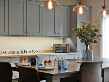 A kitchen with a wooden table and chairs plates with eggs mugs glasses a vase with green foliage hanging light fixtures and grey cabinets in the background at Bradacar in Seahouses