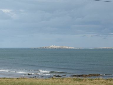 A coastal landscape with grassy shore and rocky beach looking out to a small island with a building on it at Bradacar in Seahouses