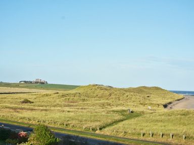 A coastal landscape with grassy dunes a road a bench and a castle in the distance at Bradacar in Seahouses