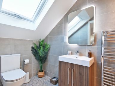 A bathroom with a toilet a sink with a wooden cabinet a lit mirror a potted plant and a skylight window at Bradacar in Seahouses