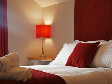 A bedroom with white bed linens and red pillows red lampshade on bedside table at Bradacar in Seahouses