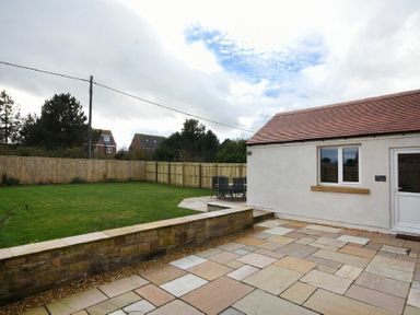 A paved patio area with a low stone wall leading to a grassy lawn and a house with white walls and a tiled roof at Bradacar in Seahouses