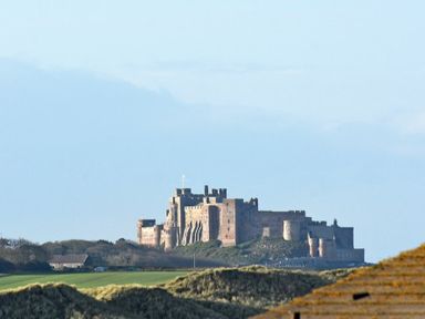 A large stone castle on a grassy hill with trees and buildings nearby at Castle View at the Viking in Seahouses